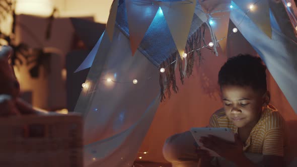 Joyous Afro-American Child Using Tablet in Cozy Teepee Decorated with Lights alt