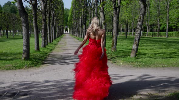 Carefree Blonde in Gorgeous Red Dress is Walking in Park in Sunny Day Enjoying Good Weather alt