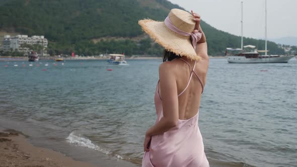 Woman in Pink Silk Dress and Straw Hat Walking on the Beach alt