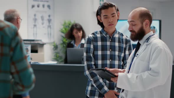 General Practitioner Talking to Asian Man About Diagnosis on Digital Tablet alt