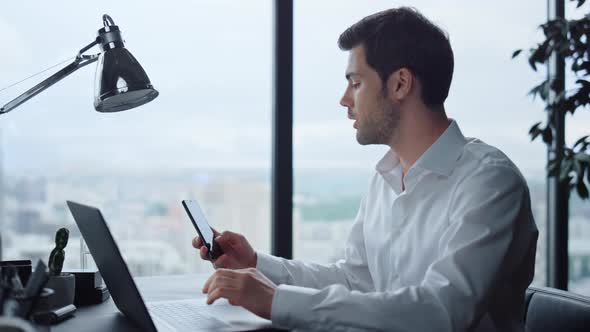 Businessman Working on Laptop at Workplace. Manager Looking at Smartphone Screen alt