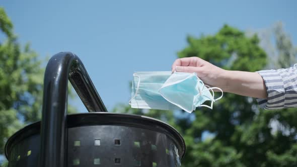 A woman's hand is throwing medical masks into a wastebasket in a park.