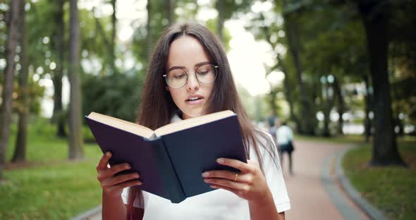 Female Student Walking and Reading Book in Park