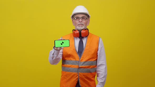 Adult man engineer in working uniform holds hand phone with greenscreen on orange background alt