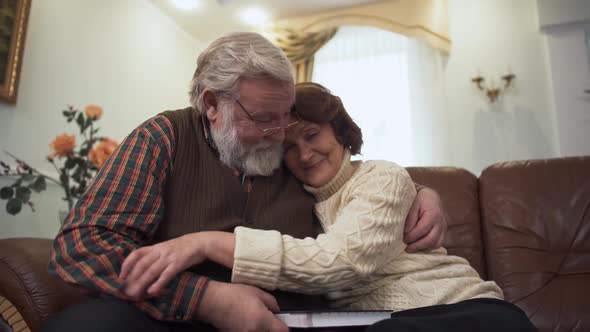 Happy Mature Couple Sitting on the Sofa in Modern Living Room at Home. Elder Man and Woman Cuddle alt