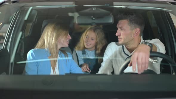 Happy Mother and Father Talking to Daughter Sitting in New Car in Dealership alt