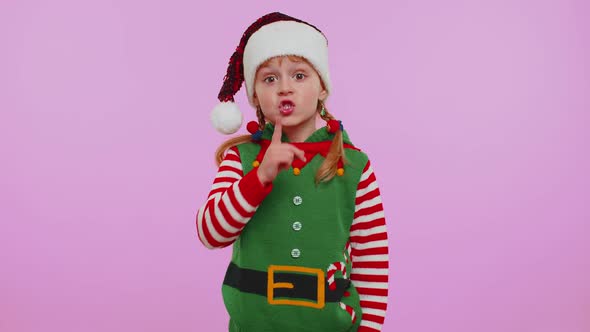 Child Girl in Christmas Elf Costume Presses Index Finger to Lips Makes Silence Gesture Sign Secret alt
