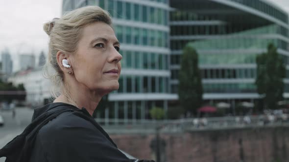 Aged Woman in Earphones Taking Break After Run at Urban Area alt