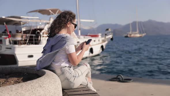 Pretty girl using smartphone while sitting at the marina. Anchored Yacht at the background alt