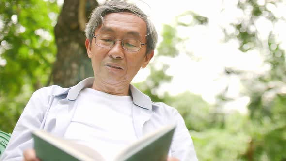 An old Asian man sits comfortably reading a book in a green park. alt