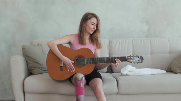 Caucasian woman with prosthetic leg is sitting on sofa and playing guitar
