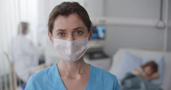 Young Nurse in Safety Mask Looking at Camera Standing in Hospital Ward with Sick Kid Lying in Bed alt