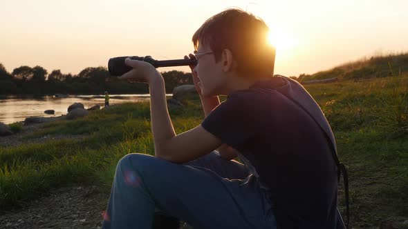 Teen Boy Sits on River Bank and Looks Into Telescope at Sunset