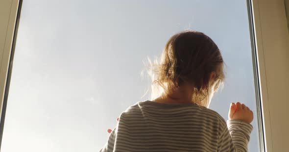 Small Child is Standing Facing the Window Looking Out at the Blue Sky in Rays of Setting Sun and