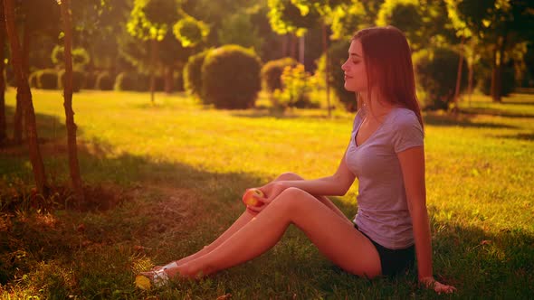 Cheerful Female Holds Fruit Sits on the Green Grass alt