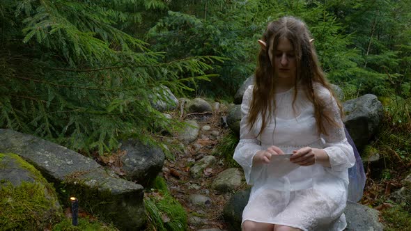 An Elf Girl in a White Dress Opens an Envelope with Letters in the Forest alt