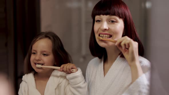 Mom and Her Cute Girl in White Bathrobe Brushing Teeth Together Standing Near the Mirror alt