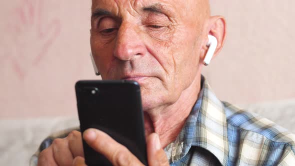 peppy caucasian pensioner 70-79 in wireless headphones uses a smartphone. an elderly man spends leis alt