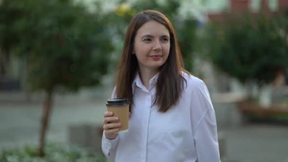 A Woman in a Strict White Shirt Drinks Coffee While Walking Along a City Street