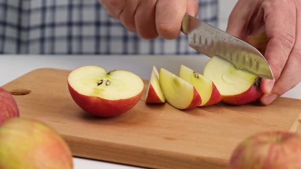 Male hands cut red apple in pieces on cutting board with knife.