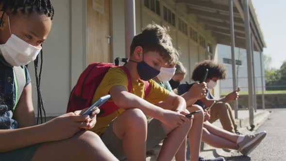 Group of kids wearing face masks using smartphones while sitting together alt