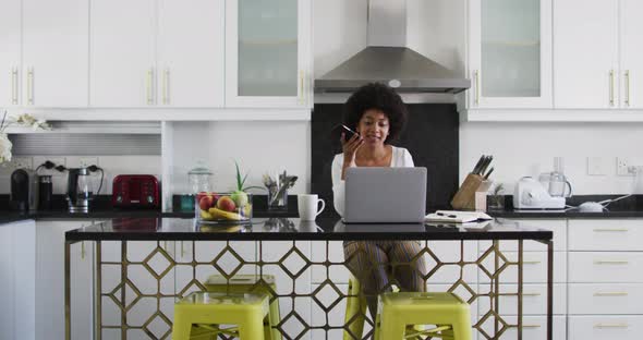 African american woman talking on smartphone and using laptop in the kitchen while working from home alt