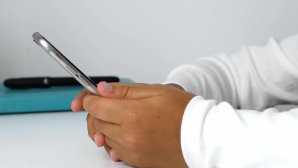 A child girl holds a phone in her hands, sits at the table, and does her homework. alt