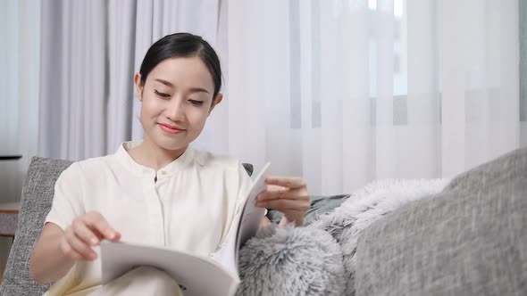 Beautiful smiling woman reading a book and sitting on the sofa in the living  alt