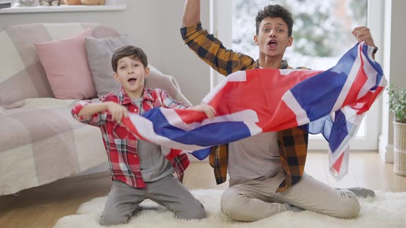 Happy Middle Eastern Teenager and Boy Rejoicing Soccer Team Victory Shaking British Flag Watching TV alt