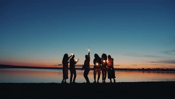 Group of Young People Friends Dancing on Lake Shore with Sparklers Late in Evening alt