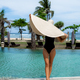 A woman in a black swimsuit stands by a pool holding a wide-brimmed straw hat - PhotoDune Item for Sale