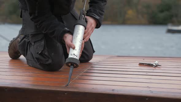 Young carpenter sealing roof planking of boat cabin with sealant gun in bleak weather. alt