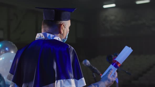 Male Lecturer r in Academic Mantle and Hat in Medical Mask Presents Diplomas to Graduates Near alt