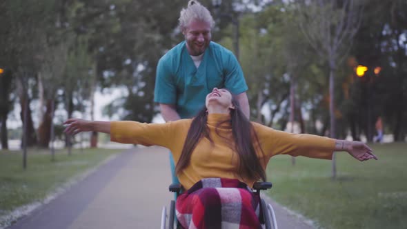 Portrait of Cheerful Paralyzed Woman with Stretched Hands Laughing As Male Nurse Running Rolling Her alt