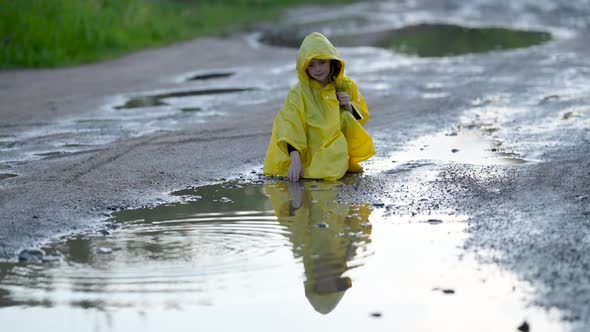 Little Cute Girl in a Yellow Raincoat Plays Near a Puddle on the road. alt
