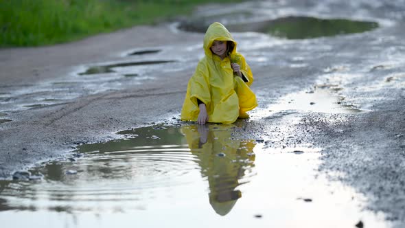 Little Cute Girl in a Yellow Raincoat Plays Near a Puddle on the road.