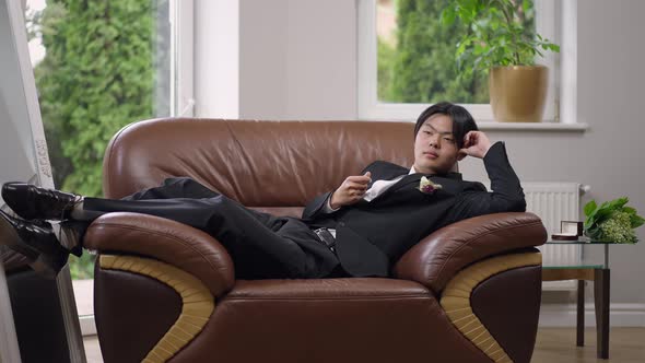Wide Shot of Happy Asian Groom Lying on Cozy Armchair Indoors Dreaming on Wedding Day alt
