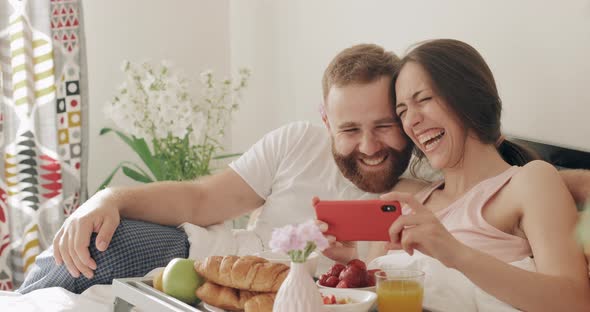 Happy Guy and Girl Watching Funny Video and Laughing in Bed. Young Romantic Family Having Breakfast alt