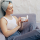 Young woman hold creme jar relaxing on couch, head wrapped in towel - PhotoDune Item for Sale
