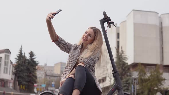Young Stylish Blonde Girl in a Light Jacket and Jeans Sits on an Electric Scooter and Takes a Selfie alt