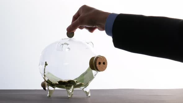 Saving Money - a Businessman Hoards Cash and Throws Some Coins Into a Glass Piggy Bank