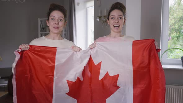 Excited Twin Sisters Hockey Fans with Canadian Flag Supporting TV Match in Living Room at Home alt