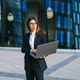 Professional woman working on a laptop outdoors, standing in front of a modern glass building - PhotoDune Item for Sale