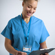 Studio Portrait Of Smiling Female Nurse Wearing Scrubs With Clipboard Against Grey Background - PhotoDune Item for Sale