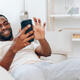 Happy African American man sitting on a black sofa, typing a message on his mobile phone He is in - PhotoDune Item for Sale