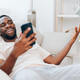 African American Man Typing on Black Smartphone while Sitting on Modern White Sofa in His Apartment - PhotoDune Item for Sale