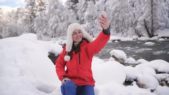 Beautiful Girl in the Mountains Near Winter River Makes Selfie on Smartphone and Smiling