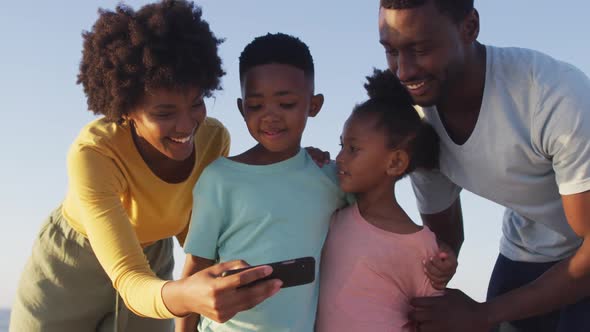Smiling african american family using smartphone and embracing on sunny beach alt