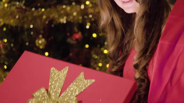 A Young Beautiful Woman Sits in a Cozy Living Room, Opens a Gift Box, Light Shines Out alt