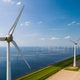 A row of wind turbines stands tall next to a serene body of water, their blades spinning gracefully - PhotoDune Item for Sale